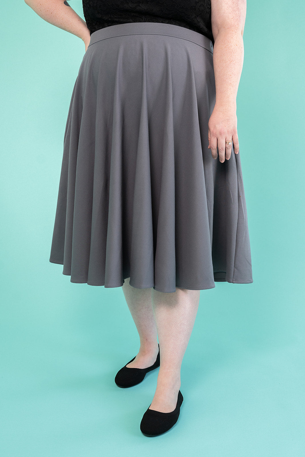 Photo of a person in a black top and grey skirt. the photo background is teal and they are wearing black flat shoes.