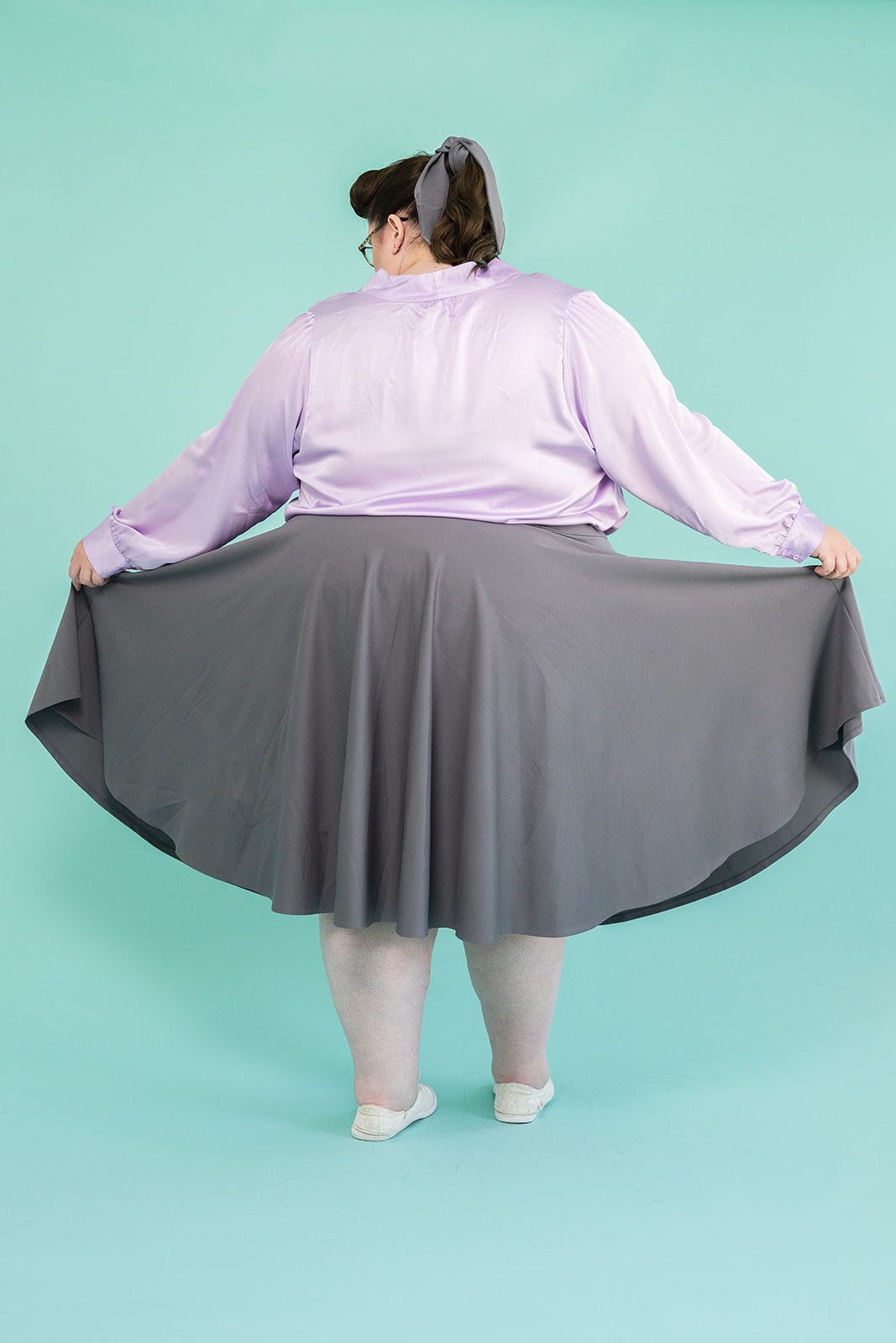 Photo of plus-size brunette woman facing away from the camera. She is holding the hem of skirt to the sides.  She is dressed in a lilac coloured blouse and grey skirt