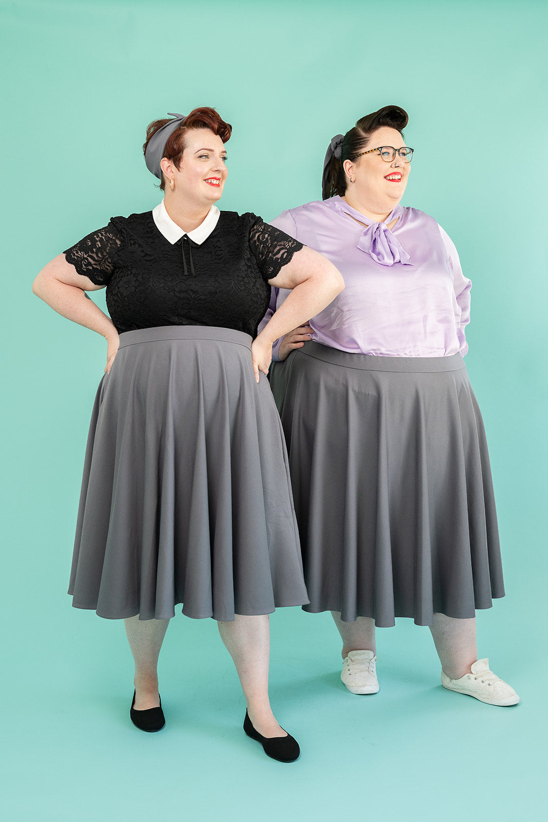Two women are modelling grey skirts. They both have brown hair. the first is wearing a black shirt with a white collar and lace sleeves. The second is wearing a lilac silk shirt with a bow at the neckline. 