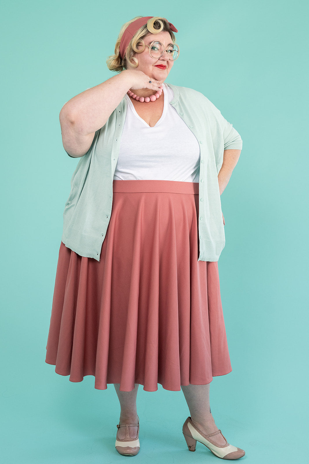 A plus-size blonde woman is looking at the camera and playfully smiling. She is wearing a light blue cardigan, a white tshirt and a pink skirt