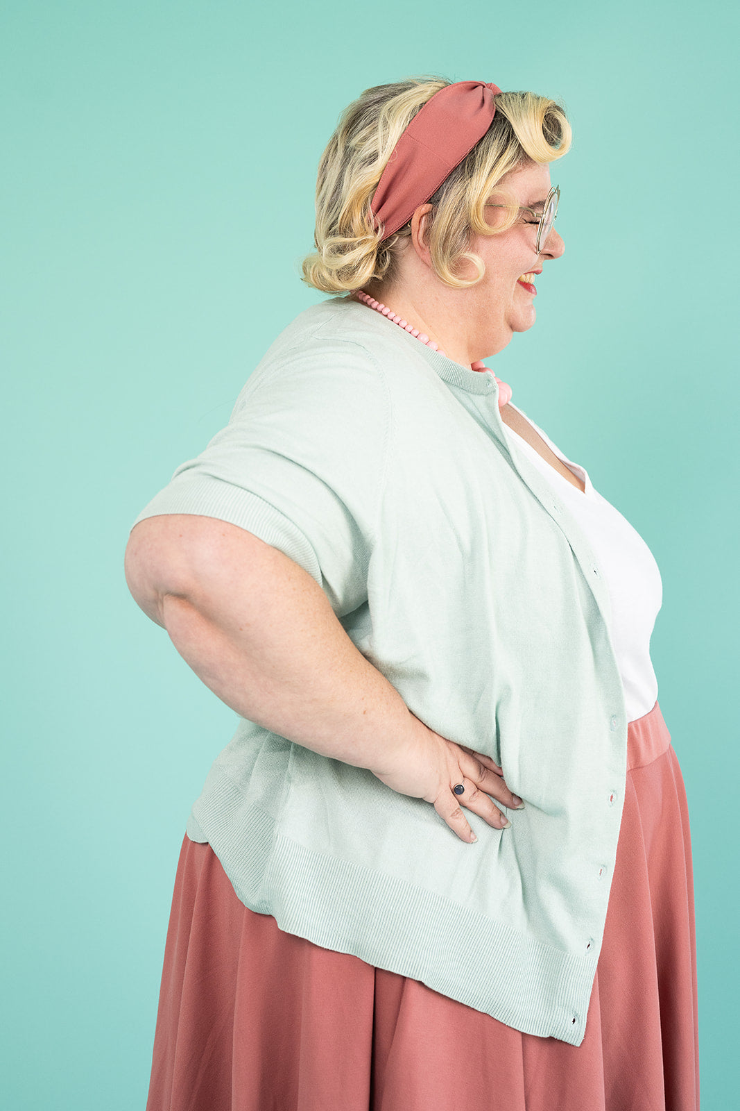 A plus-size blonde woman is standing with her hands on her hips and smiling. She is wearing a light blue cardigan, a white tshirt and a pink skirt