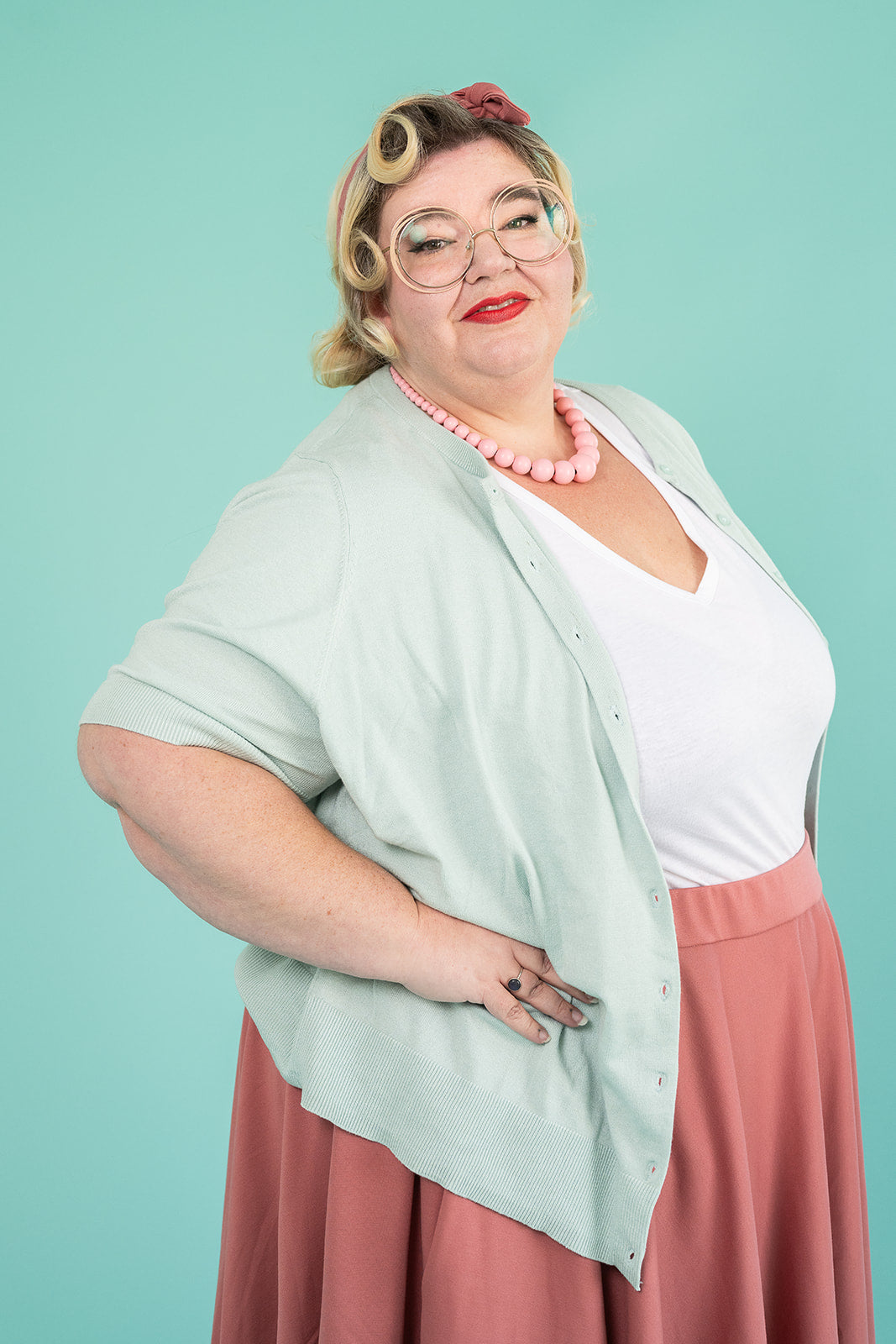 A plus-size blonde woman is standing with her hands on her hips and smiling. She is wearing a light blue cardigan, a white tshirt and a pink skirt