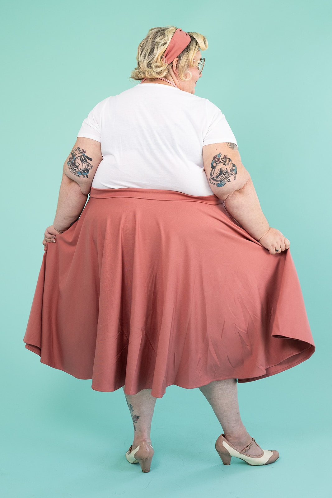 A plus-size blonde woman is facing away from the camera and showing off her skirt. She has several tattoos on her arms; she is wearing a white tshirt and a pink skirt