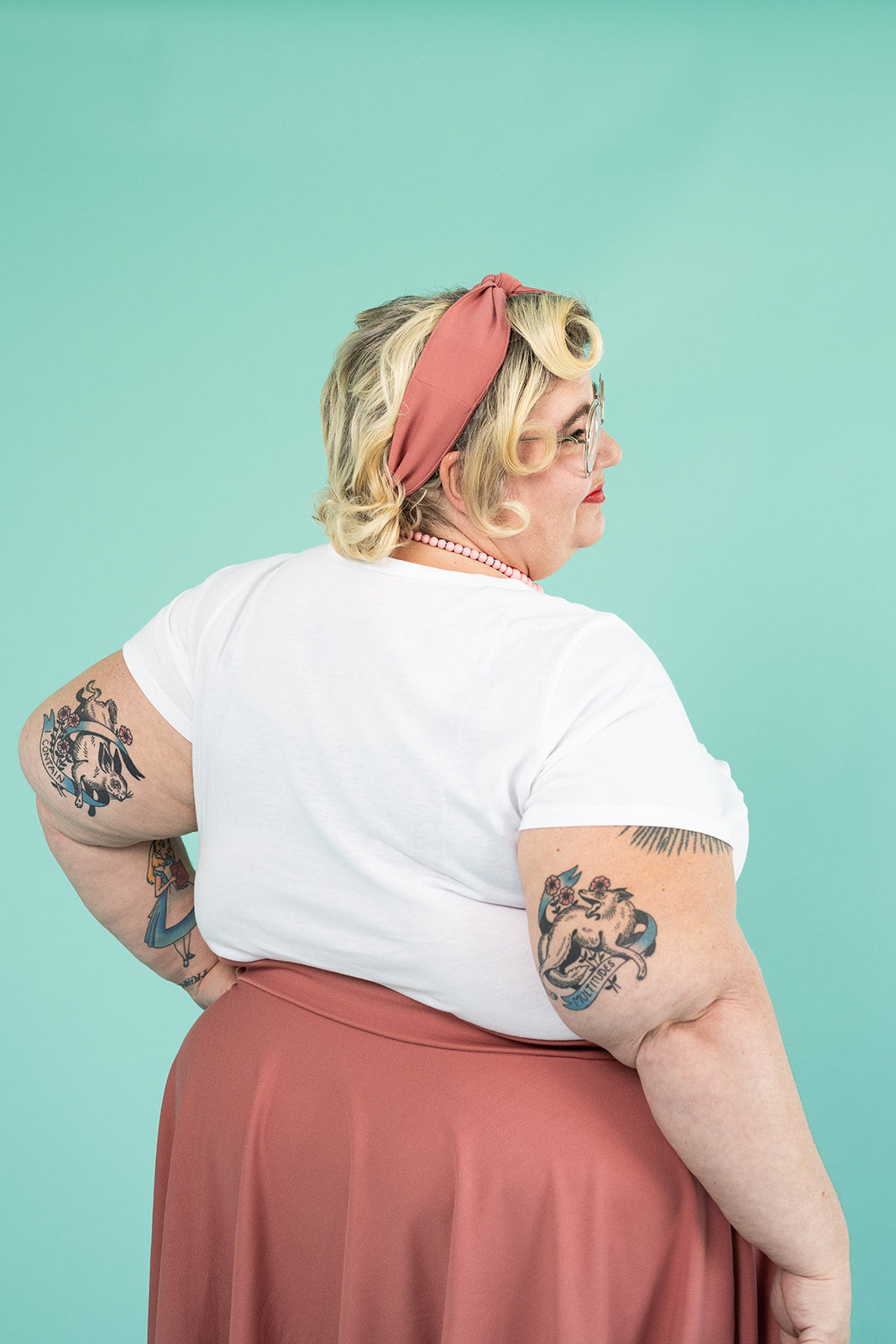 A plus-size blonde woman is facing away from the camera. She has several tattoos on her arms; she is wearing a white tshirt and a pink skirt