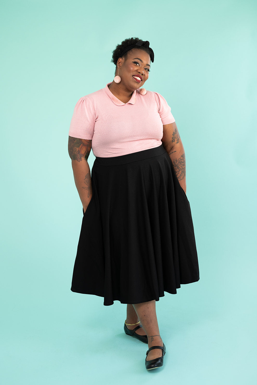 A black woman is looking at the camera and smiling. She is wearing a pink top and pompom earrings and a black skirt.