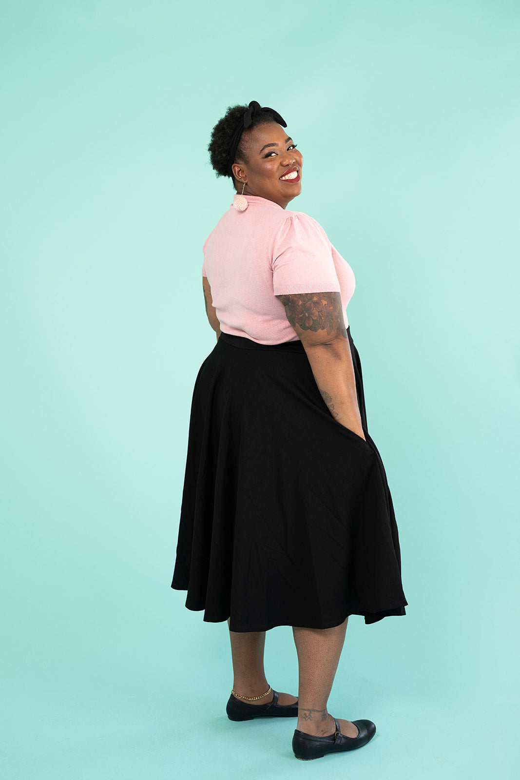 A black woman is turned away but looking at the camera and smiling. She is wearing a pink top and pompom earrings and a black skirt.