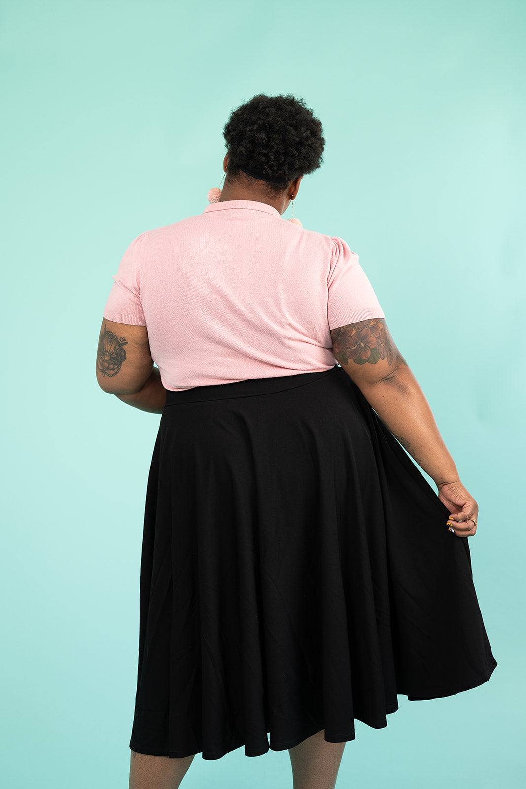 A black woman is facing away from the camera and modelling her skirt. She is wearing a pink top and pompom earrings and a black skirt.