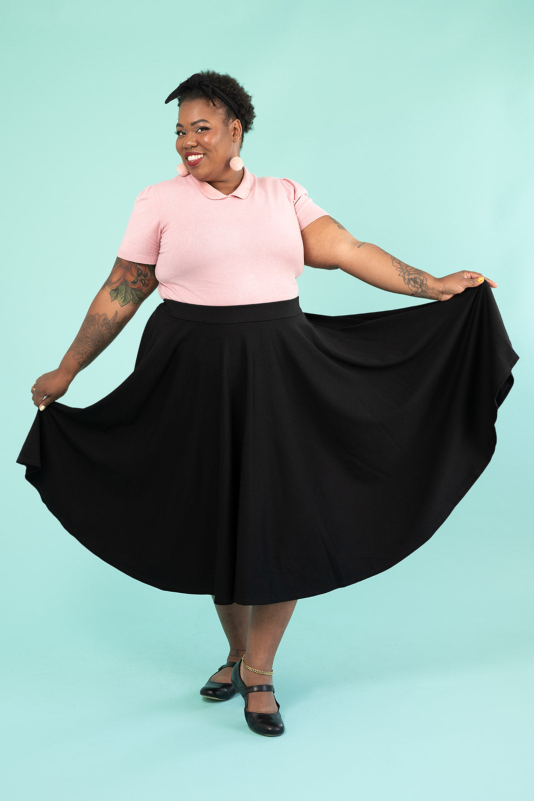 A black woman is jubilantly smiling and showing off her skirt. She is wearing a pink top and pompom earrings and a black skirt.
