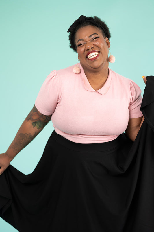 A black woman is jubilantly smiling and showing off her skirt. She is wearing a pink top and pompom earrings and a black skirt.