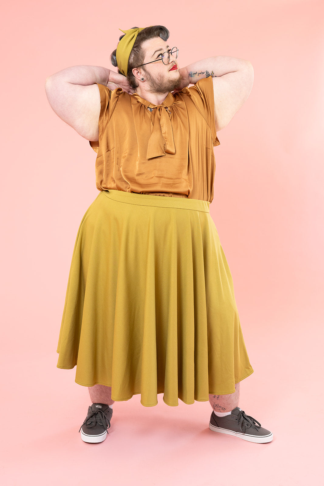 A gender non-conforming person is posing with their arms by their face.  They are wearing red lipstick and have their hair in a vintage-inspired updo. They have facial hair. They are wearing a bronze silk shirt and mustard skirt.