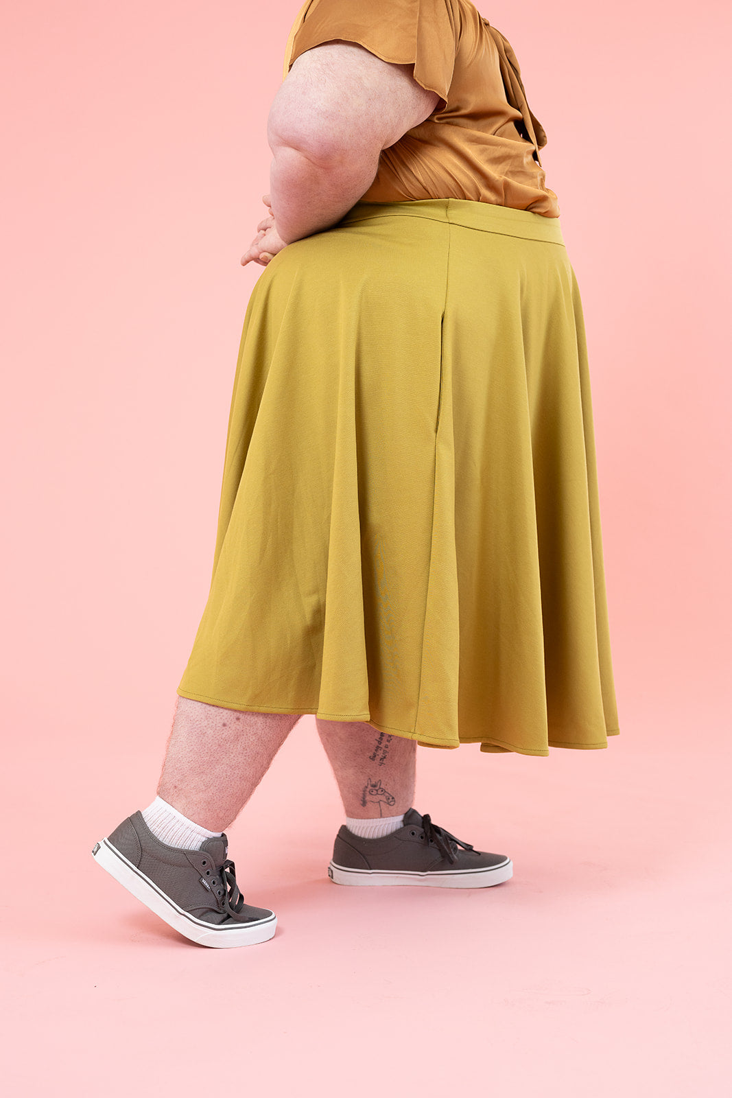 A person is shown from the waist down wearing a bronze silk shirt and a mustard colour skirt with grey sneakers. They have several tattoos. 