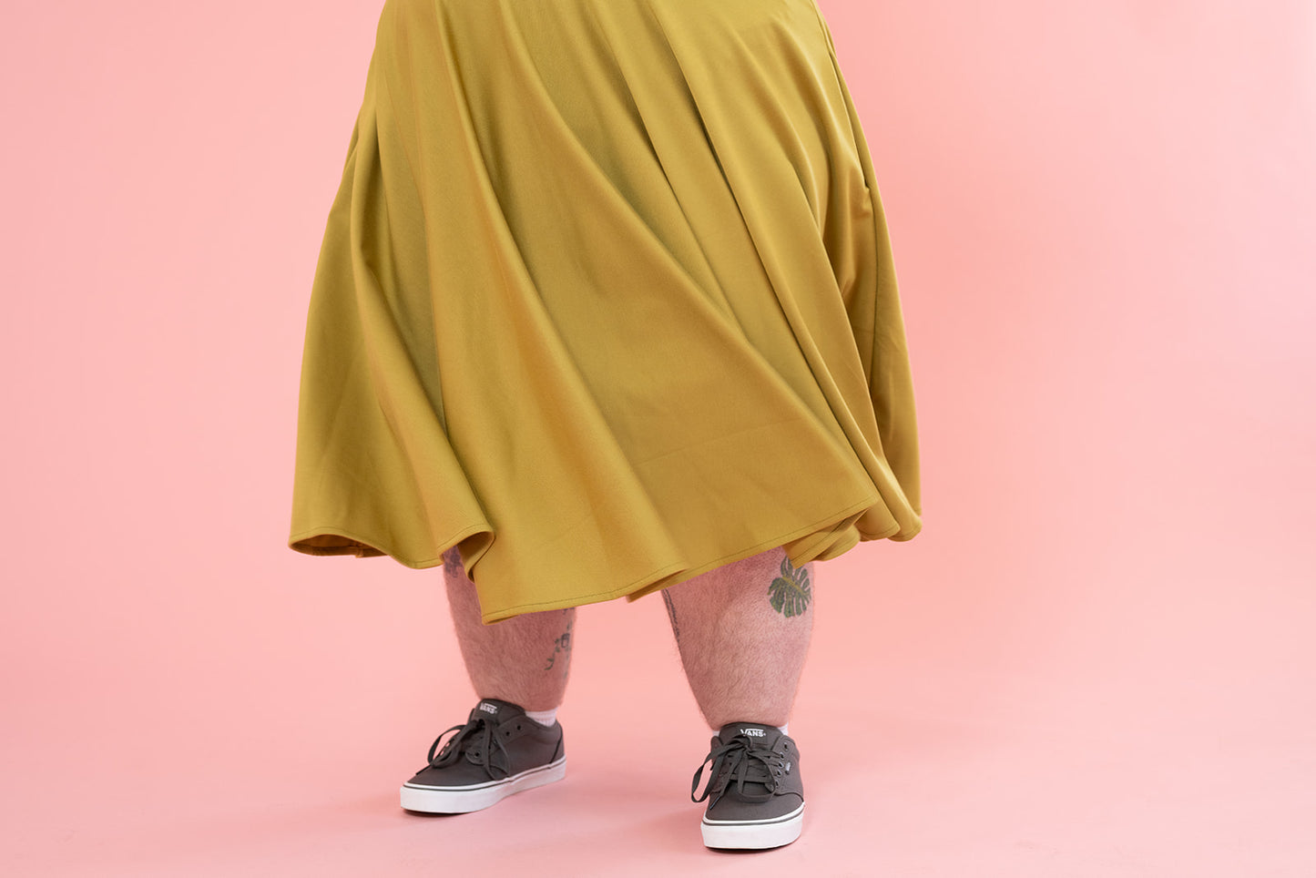 Photo of a person wearing a mustard coloured skirt. They have a few tattoos on their legs and are wearing grey sneakers.