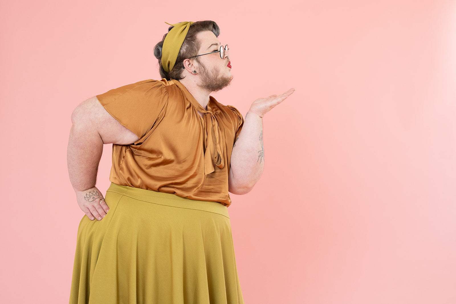 A gender non-conforming person is blowing a kiss. Their outfit is vintage-inspired with a bronze silk bow top and mustard skirt. 