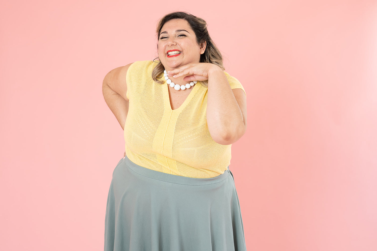 A plus-size white woman is smiling and posing for the camera; she is wearing a light yellow shirt, white beaded necklace and blue skirt. 