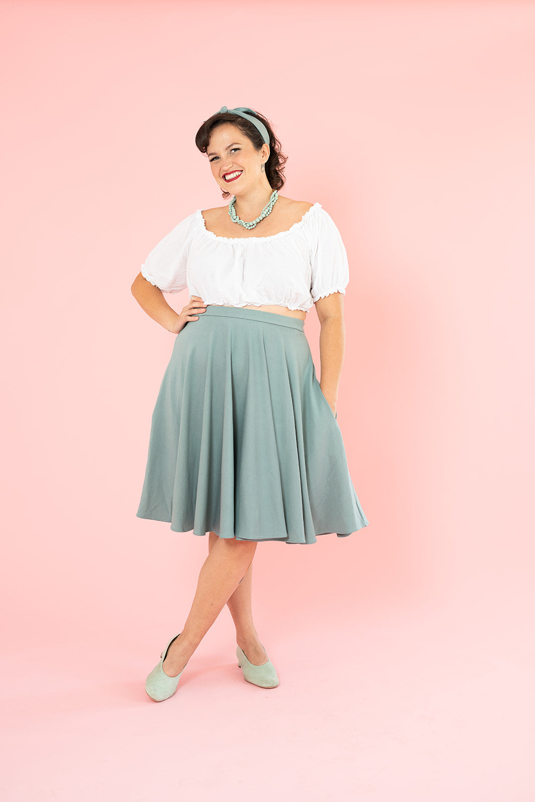 A smiling young white woman with dark brown hair wearing a white top and a flowy vintage-inspired mint skirt.
