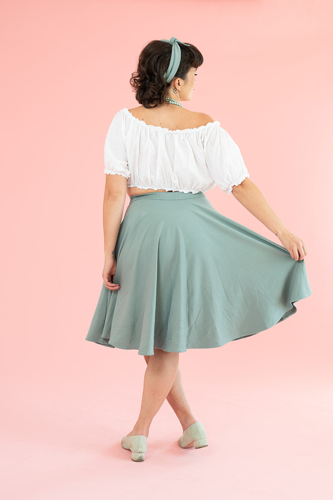 A young white woman with dark brown hair in a white top holds out the hem of a flowy vintage-inspired mint skirt.