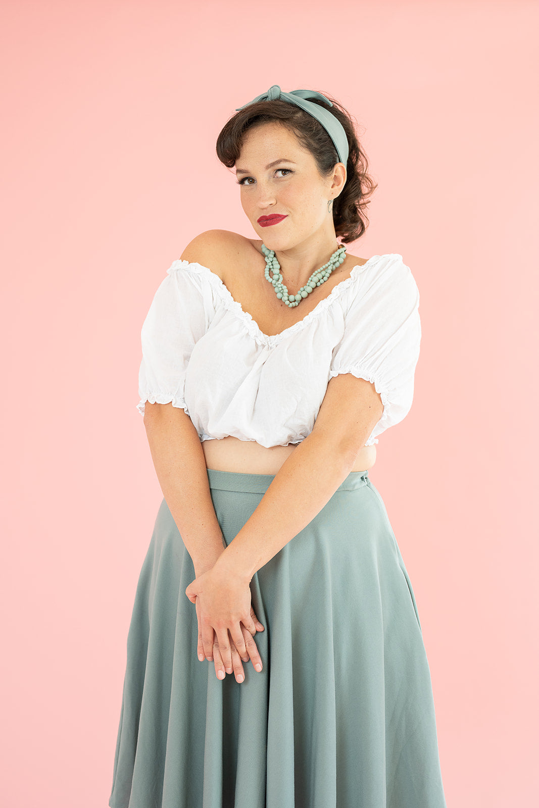 A woman with brown hair looks at the camera. She is wearing a white cropped top with off the shoulder sleeves and a blue skirt.